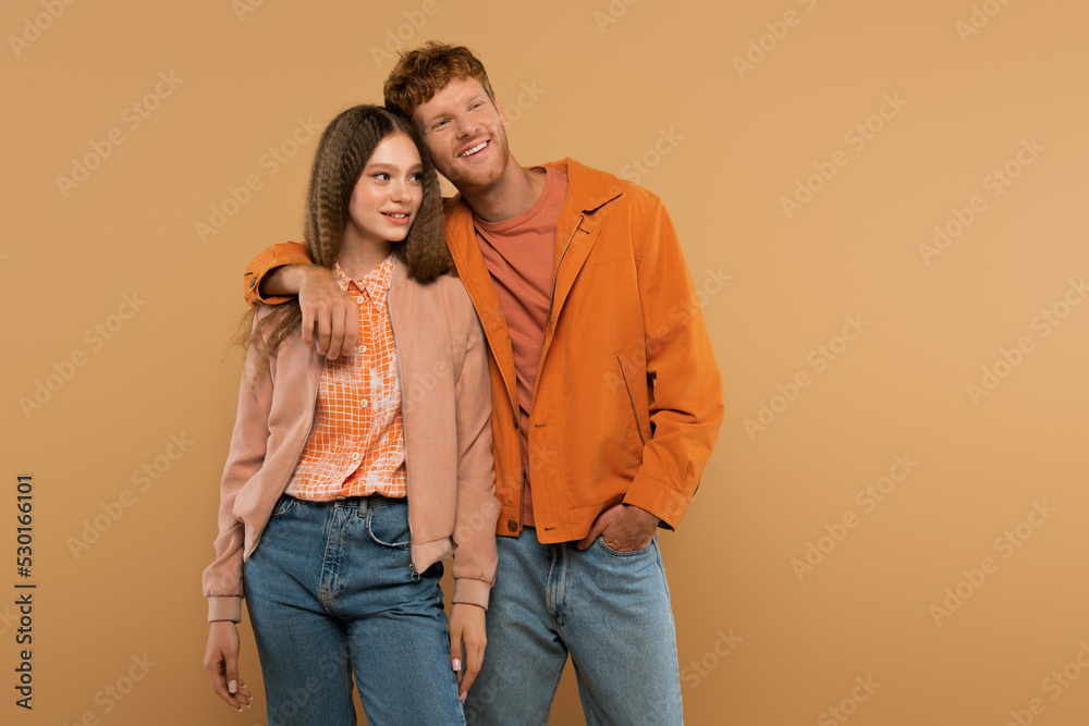 Fototapeta premium redhead young man standing with hand in pocket and hugging cheerful girlfriend isolated on beige.