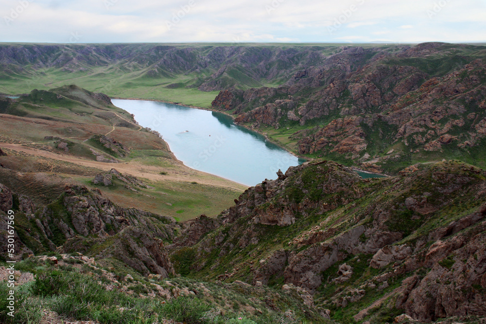 Fototapeta premium Top view of a wide river in a mountain valley, cloudy summer day, the sky is in the clouds, the mountain slopes are covered with rocks and green grass, there is sand on the river bank, Ili river, Kaza