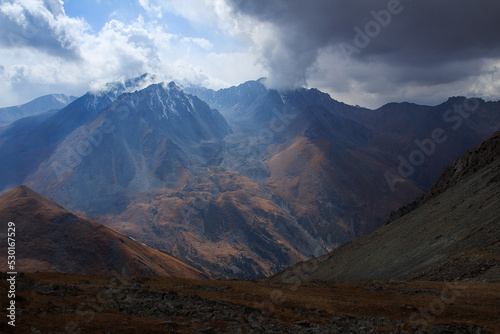 Alpine gorge with peaked mountain peaks, the slopes are covered with stones, from the side the sun illuminates the gorge, clouds in the sky hang on the mountain tops, Prokhodnoye gorge, Kazakhstan