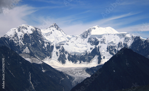 Mount Belukha with snow-capped peaks  below there is a glacier  in the foreground peaked stone mountains  a clear day  the sky is in clouds