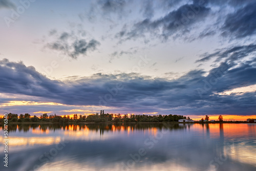 Summer sunset on Lake. Natural rural landscape, autumn September evening. Village on lakeside. Dusk rural scene. Storm cloudscape