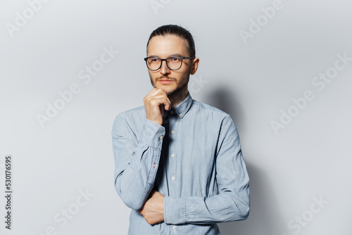 Studio portrait of young thoughtful man in blue shirt wearing eyeglasses, touching his chin on white background.