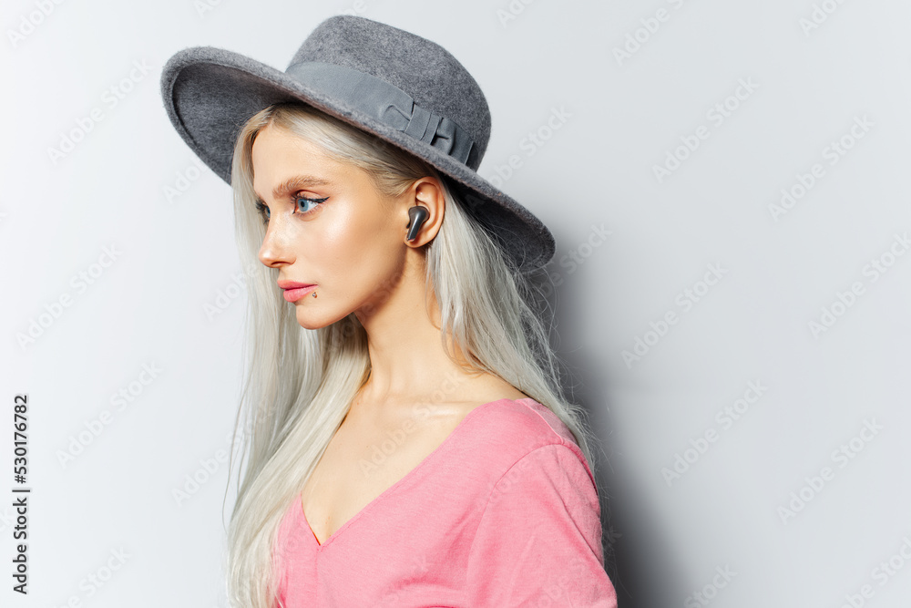 Studio side portrait of young pretty blonde girl, listen the music with wireless earbuds, wearing grey hat and pastel pink shirt on white background. Studio side portrait of young pretty blonde girl, listen the music with wireless earbuds, wearing grey hat and pastel pink shirt on white background.