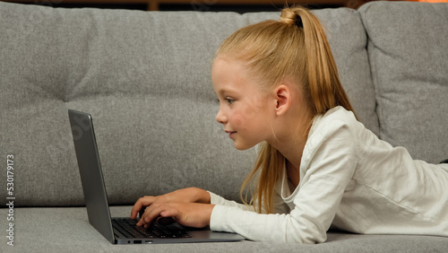 Carefree little kid pretty blonde girl child daughter using laptop lying on grey sofa surfing internet chatting with classmates playing games studying online at home. Children tech addiction concept photo