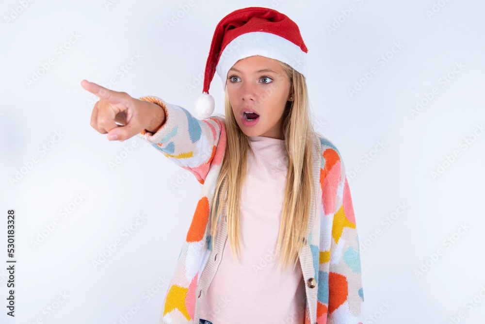 little kid girl with Christmas hat wearing yarn jacket over white background Pointing with finger surprised ahead, open mouth amazed expression, something on the front. little kid girl with Christmas hat wearing yarn jacket over white background Pointing with finger surprised ahead, open mouth amazed expression, something on the front.