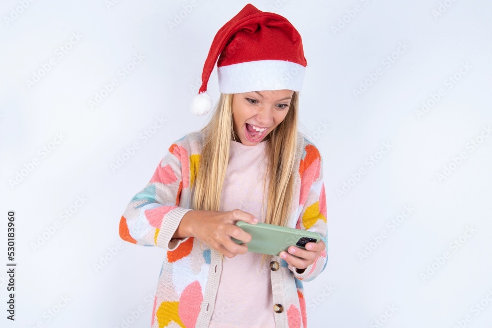 Portrait of an excited little kid girl with Christmas hat wearing yarn jacket over white background playing games on mobile phone. Portrait of an excited little kid girl with Christmas hat wearing yarn jacket over white background playing games on mobile phone.