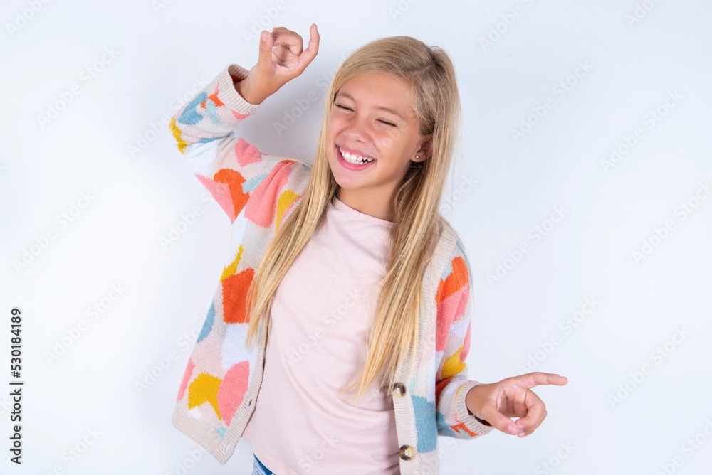 Photo of upbeat little kid girl wearing colorful yarn jacket over white background has fun and dances carefree wear being in perfect mood makes movements. Spends free time on disco party Photo of upbeat little kid girl wearing colorful yarn jacket over white background has fun and dances carefree wear being in perfect mood makes movements. Spends free time on disco party
