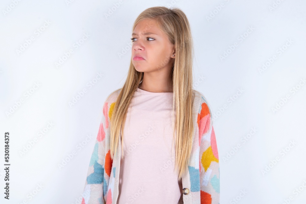 Dissatisfied little kid girl wearing colorful yarn jacket over white background purses lips and has unhappy expression looks away stands offended. Depressed frustrated model. Dissatisfied little kid girl wearing colorful yarn jacket over white background purses lips and has unhappy expression looks away stands offended. Depressed frustrated model.