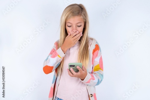 little kid girl wearing colorful yarn jacket over white background being deeply surprised, stares at smartphone display, reads shocking news on website, Omg, its horrible!
