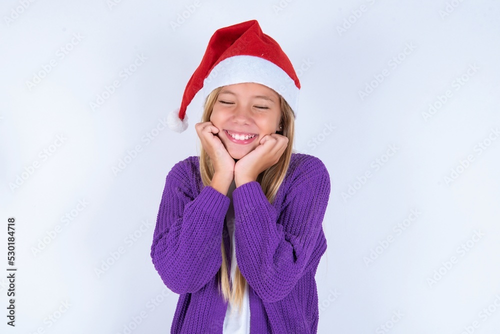 little kid girl with Christmas hat wearing yarn jacket over white background grins joyfully, imagines something pleasant, copy space. Pleasant emotions concept. little kid girl with Christmas hat wearing yarn jacket over white background grins joyfully, imagines something pleasant, copy space. Pleasant emotions concept.