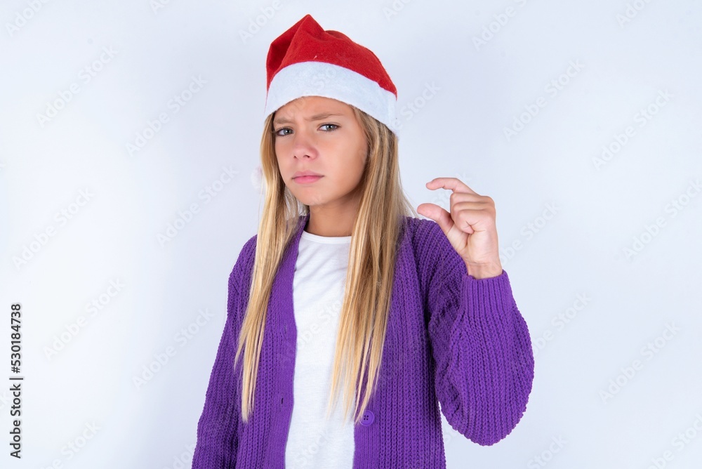 Displeased little kid girl with Christmas hat wearing yarn jacket over white background shapes little hand sign demonstrates something not very big. Body language concept.