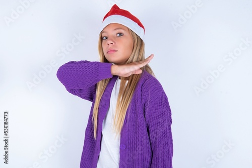 little kid girl with Christmas hat wearing yarn jacket over white background cutting throat with hand as knife, threaten aggression with furious violence. photo