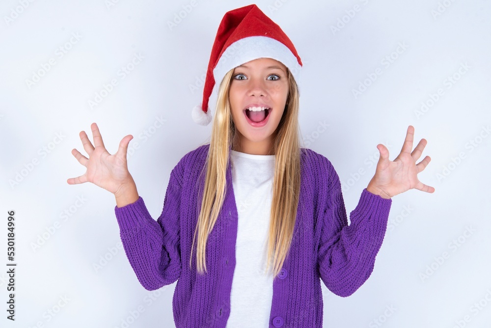 Delighted positive little kid girl with Christmas hat wearing yarn jacket over white background opens mouth and arms palms up after having great result Delighted positive little kid girl with Christmas hat wearing yarn jacket over white background opens mouth and arms palms up after having great result