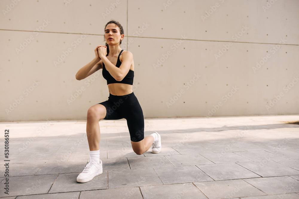 Attractive young sporty woman staying at street and doing luges. Have workout outdoors on the wall background wearing black sportswear. Concept of health life, exercises Attractive young sporty woman staying at street and doing luges. Have workout outdoors on the wall background wearing black sportswear. Concept of health life, exercises