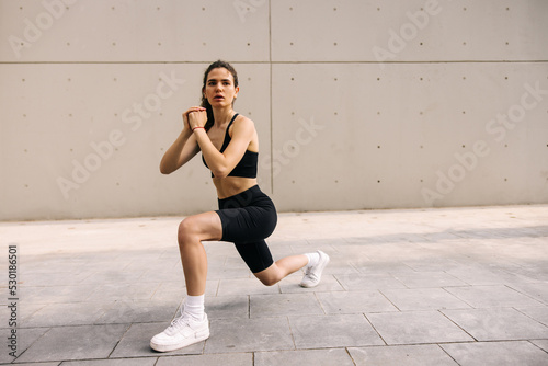Full-length view of caucasian dark-haired woman doing exercise at street, looking away. Staying in lunges pose on wall background. Workout outdoors concept © Look!