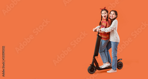 Portrait of cute twin girls with kick scooter on orange background with space for text
