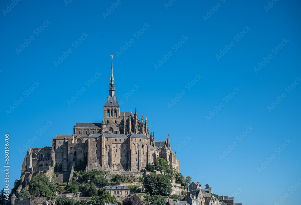 Mont St. Michel, Normandy, France - July 8, 2022: Historic brown stone abbey part with church on top of rock against blue sky. Some green foliage on flank of hill.