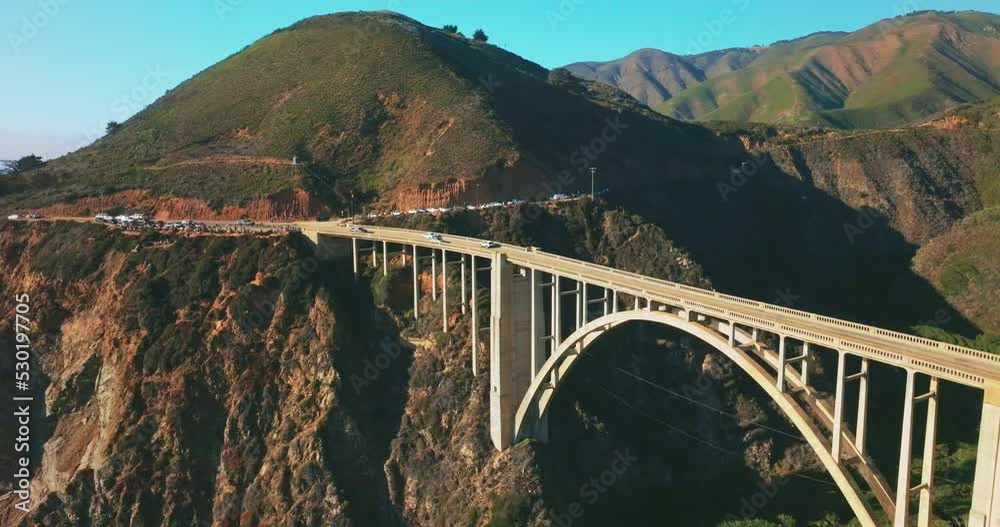 Busy road around the rugged mountain passing into the beautiful arched bridge over the gap in the rocks. Blue skies at backdrop. Top view. Busy road around the rugged mountain passing into the beautiful arched bridge over the gap in the rocks. Blue skies at backdrop. Top view.