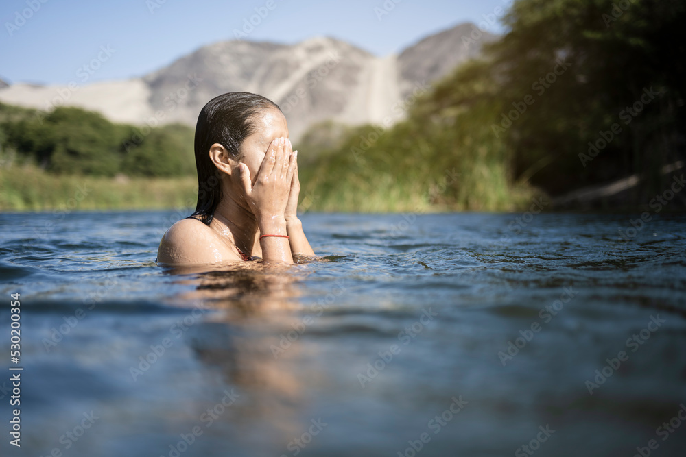 Fototapeta premium Beautiful young latin woman bathes in a lake enjoying a summer day. It stands at the bottom of the shoulders in the water. Her hands are raised above the water.