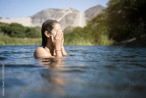 Beautiful young latin woman bathes in a lake enjoying a summer day. It stands at the bottom of the shoulders in the water. Her hands are raised above the water.