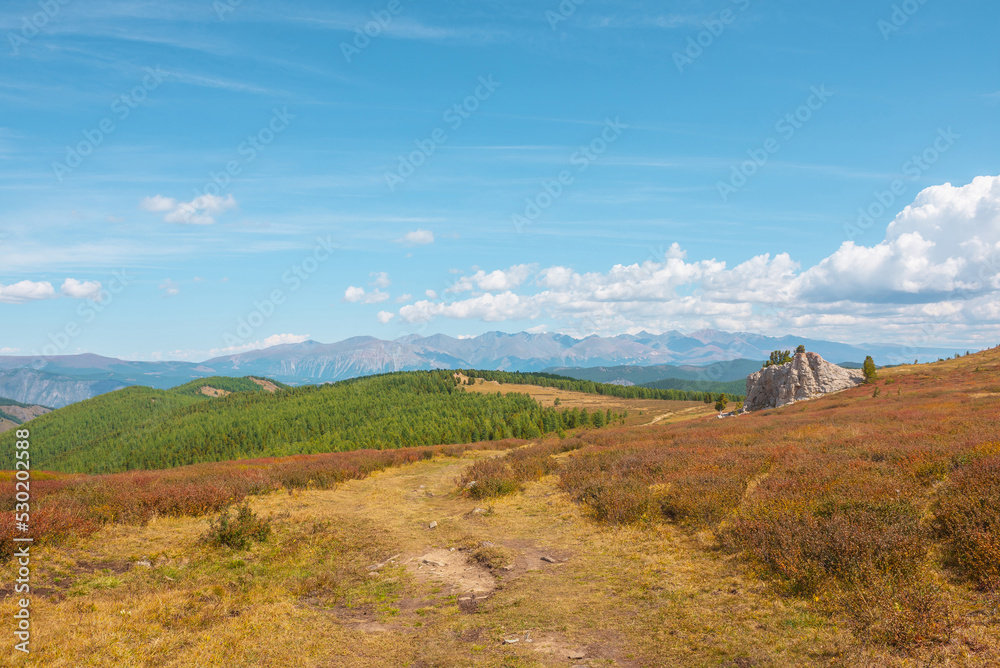 Atmospheric autumn landscape with big rock on hill with view to sunlit mountain vastness and high mountain range line on horizon under clouds. Impressive mountain scenery in vivid fading autumn colors Atmospheric autumn landscape with big rock on hill with view to sunlit mountain vastness and high mountain range line on horizon under clouds. Impressive mountain scenery in vivid fading autumn colors