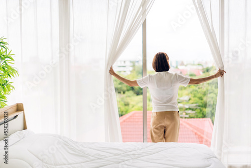 Smiling of happy young beautiful pretty asian woman waking up and opening window curtains.Girl feeling comfortable and relaxed breathing fresh air in morning in bedroom at home