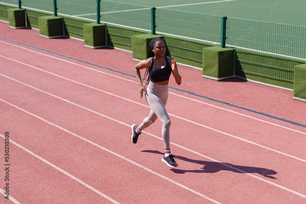 Young black sportswoman enjoys running on red track around stadium to improve technique and increase endurance. African American woman with long braids prepares for upcoming marathon on hot summer day