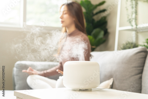 Air humidifier, calm blurred woman, girl sitting on couch lotus pose put hands practice meditation do yoga exercise at home. Aromatherapy steam scent from essential oil diffuser in living room at home