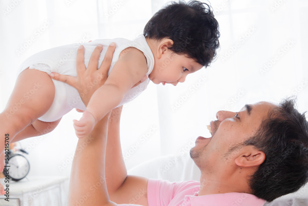 Selective focus asian father play and tease with newborn baby in bedroom. Dad holding lifting toddler on bed with fun and tender. Adorable little boy having fun with daddy togerther at home. Selective focus asian father play and tease with newborn baby in bedroom. Dad holding lifting toddler on bed with fun and tender. Adorable little boy having fun with daddy togerther at home.