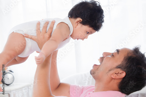 Selective focus asian father play and tease with newborn baby in bedroom. Dad holding lifting toddler on bed with fun and tender. Adorable little boy having fun with daddy togerther at home. photo