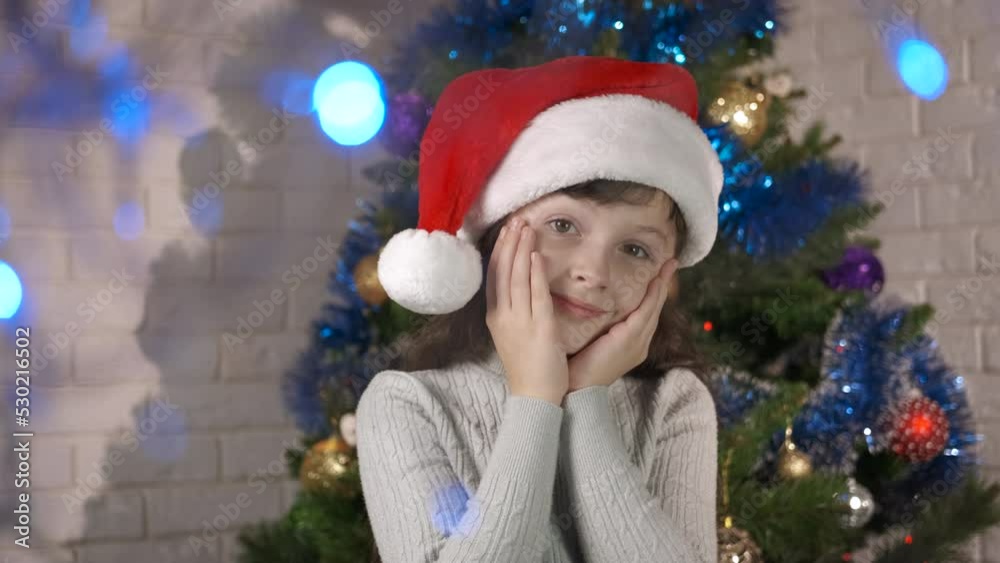 Relaxation in empty decorated room. A cute emotional cute girl in Santa hat stay in the room by Christmas tree. Relaxation in empty decorated room. A cute emotional cute girl in Santa hat stay in the room by Christmas tree.
