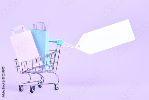 Shopping cart full of bags with a blank label on a pastel lilac background. Minimalist design with copy space. Concepts: market deals, seasonal sales and discounts, black friday advertising.