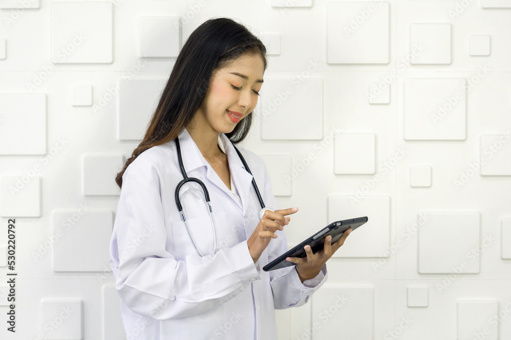 Young asian doctor in white gown and stethoscope typing on tablet computer, stand in front of white rectangle pattern ceramic wall. Young asian doctor in white gown and stethoscope typing on tablet computer, stand in front of white rectangle pattern ceramic wall.