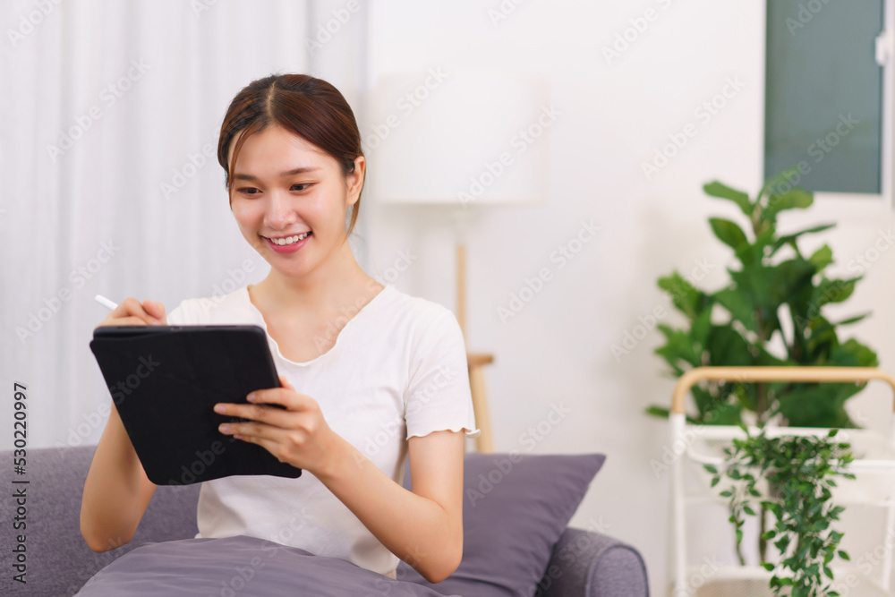 Lifestyle in living room concept, Young Asian woman writing data on tablet while sitting on couch Lifestyle in living room concept, Young Asian woman writing data on tablet while sitting on couch