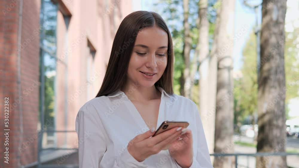 Calm beautiful woman standing outdoor using smartphone near office place. Lovely girl brunette scrolling mobile gadget, browsing internet, social media relaxing under tree on city street. Calm beautiful woman standing outdoor using smartphone near office place. Lovely girl brunette scrolling mobile gadget, browsing internet, social media relaxing under tree on city street.