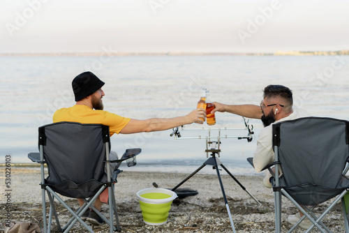 Two fishermen sitting together with beer while fishing on the lake at the summer morning. leisure and people
