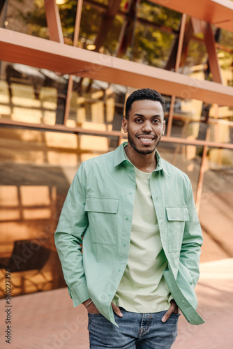Smiling young african guy looks at camera, keeps his hands in jeans pockets standing on street. Brunette man with stubble wears shirt. Happy weekend concept.