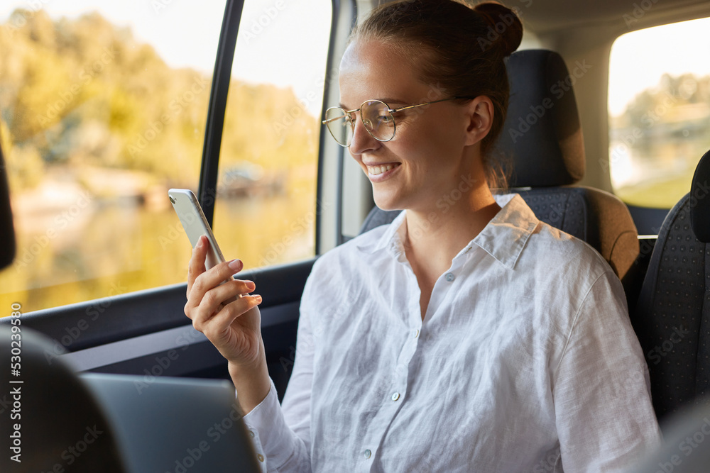 Portrait of happy optimistic woman in glasses and with bun hairstyle using laptop and mobile phone while sitting in the car, reading message on phone or checking social networks.