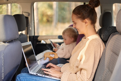 Profile portrait of smiling delighted woman working on portable computer while sitting with her baby daughter in safety chair on backseat of the automobile.
