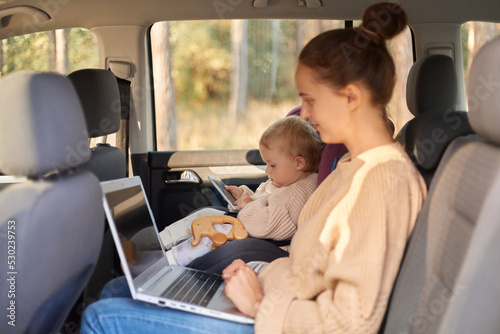Profile portrait of attractive beautiful young adult woman working on laptop while sitting with her infant daughter in safety chair on backseat of the her vehicle.