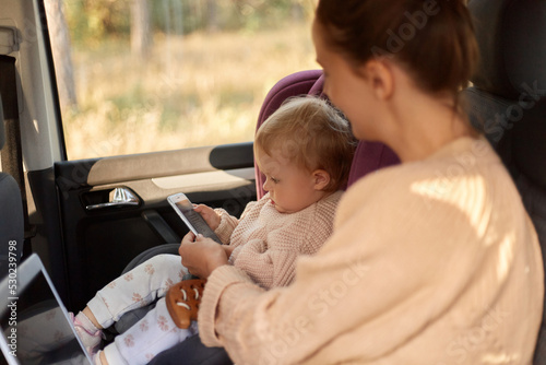 Side view portrait of busy woman working on laptop and playing with her toddle daughter while sitting in safety chair on backseat of the car, showing to her kid how to use phone. photo