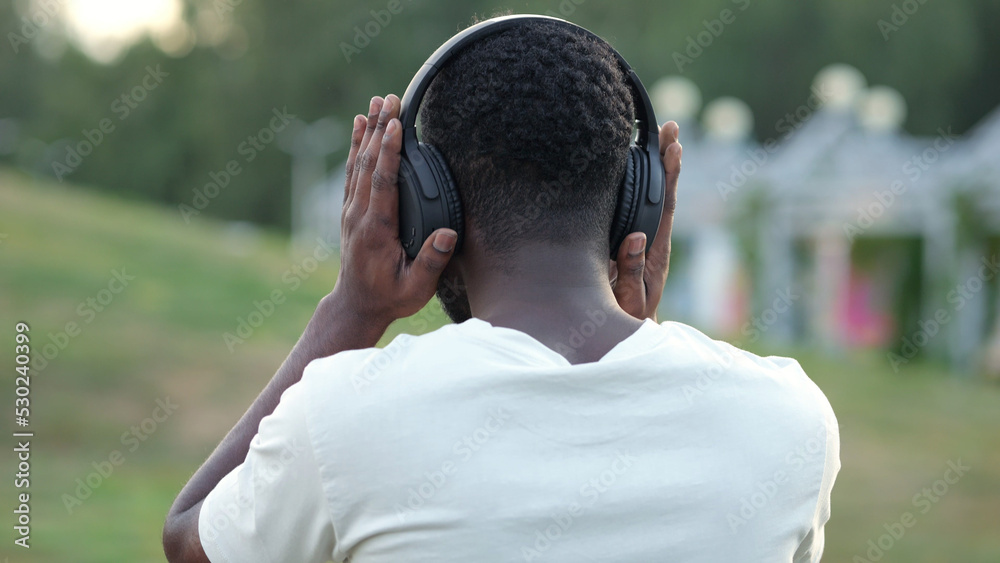 Black man putting on headphones walks with motivation in green park. African American tourist goes with confident posture continuing successful day