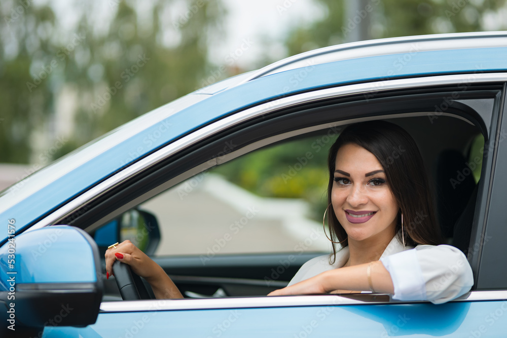 Adult black-haired woman driver sits in blue car. Stylish businesswoman enjoys successful life smiling. Happy lady grimaces in leather cabin closeup