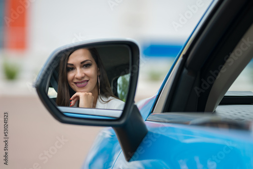 Woman with make-up looks at own reflection in side rear view mirror with satisfied expression. Successful driver enjoys morning sitting in blue car