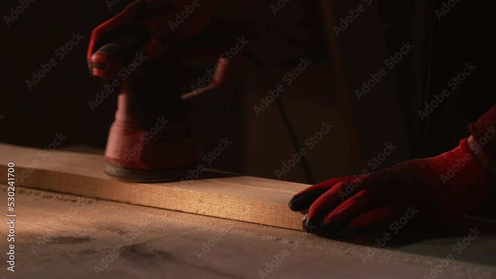 Joinery work. Man hobby. DIY carpentry. Carpenter hands in gloves sanding wood plank using grind machine tool at dark workplace with sawdust.