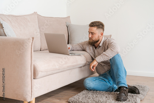 Young attractive, smiling bearded guy browsing his laptop while sitting at home on a cozy beige sofa at home, in casual clothes on a day off © Екатерина Переславце