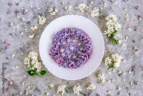 Served plate with purple and white lilac flowers on gray concrete background, top view. Spring season concept with minimal design.