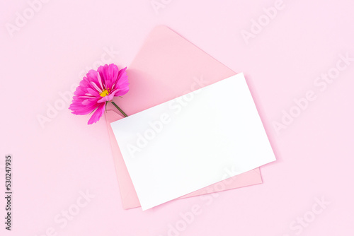 White blank card, pink envelope and flowers on pink background. Minimal style. Top view Flat lay Mockup