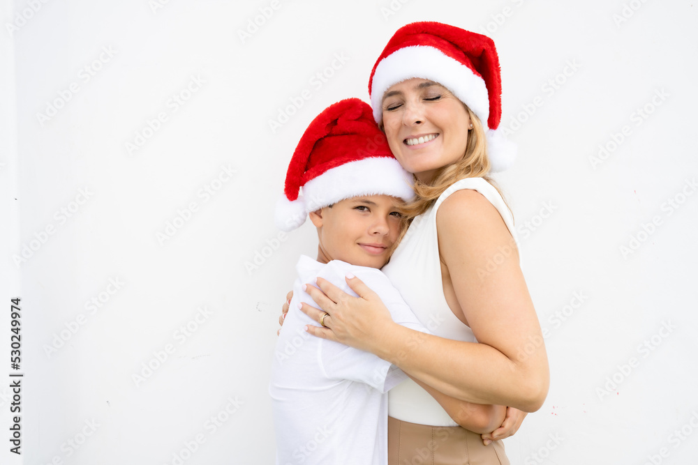 Mother and son in Santa hats smiling enjoying Christmas season on white wall. Mother and son in Santa hats smiling enjoying Christmas season on white wall.