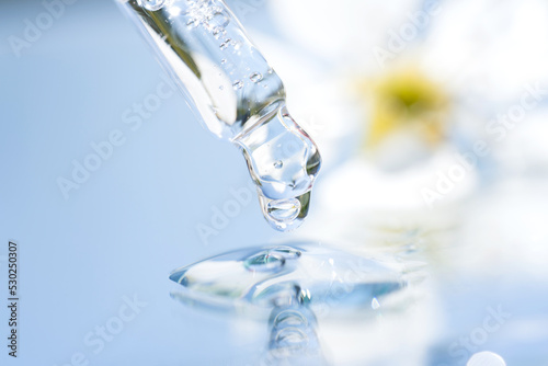 Dripping drop of transparent liquid with glass pipette against light blue background with flowers. Texture of face serum, herbal essence or natural cosmetic oil. Selective focus